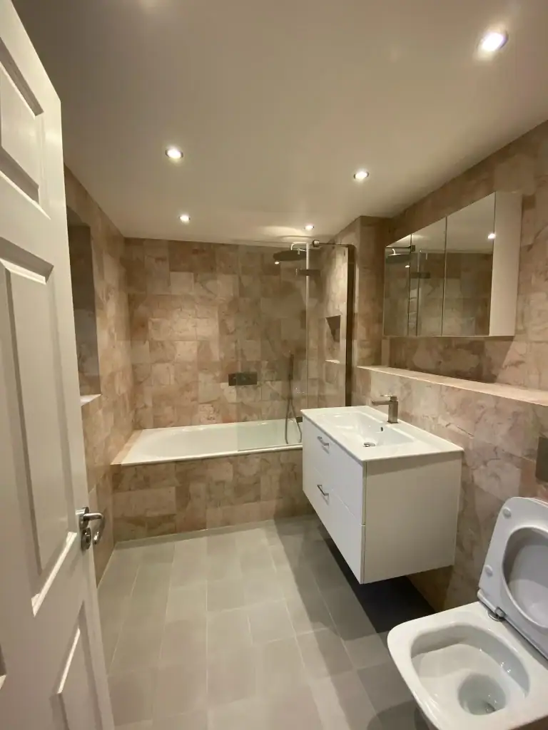 Close-up of curved white bathtub integrated with overhead rainfall shower, chrome mixer tap, and surrounding beige terracotta tiles in a modern Dulwich bathroom