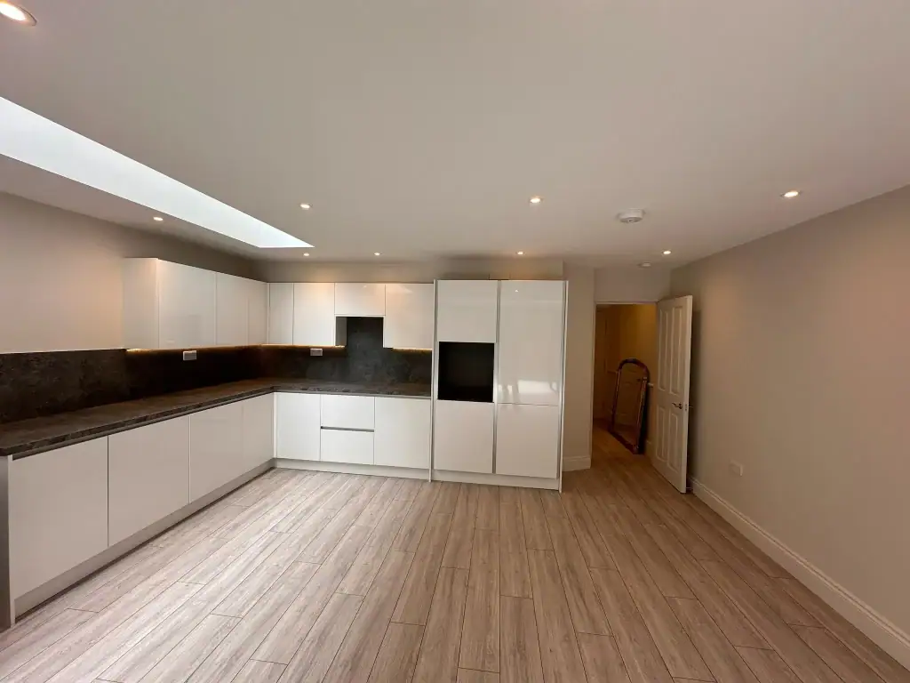 Spacious Balham kitchen with glossy white wall and base cabinets, black granite splashback, integrated ovens, utility door, recessed spotlights, and neutral walls under a linear skylight