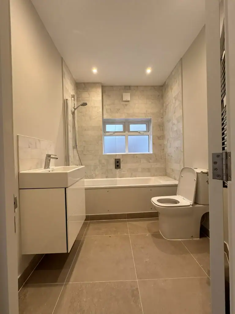 Bright Balham bathroom with white marble wall tiles, freestanding white vanity unit, integrated bathtub with overhead shower, sleek white toilet, grey porcelain floor tiles, and frosted windows
