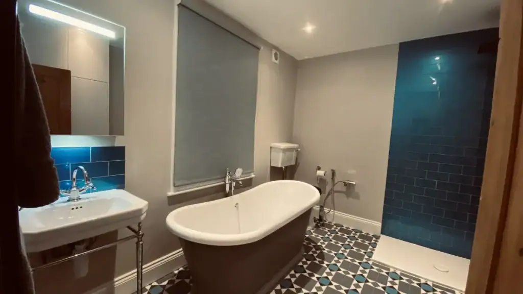 Elegant white freestanding bathtub on gray-and-blue patterned Victorian-style floor tiles, with chrome lever taps and chimney breast shelving in a Dulwich luxury renovation.