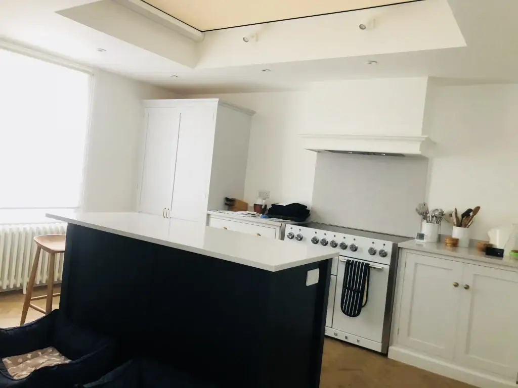 Wide shot of matte black island unit with white quartz top, wooden bar stools, pendant space, and under-cabinet drawers in a luxurious Kensington kitchen