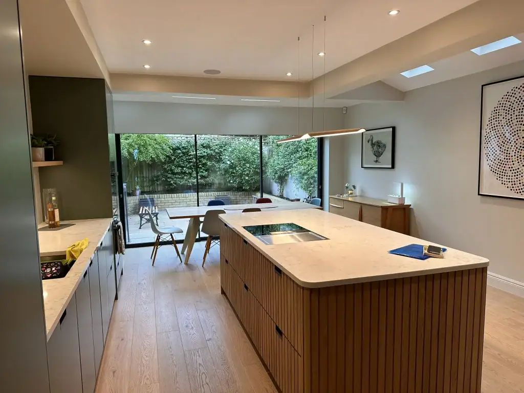 Wide shot highlighting vertical oak panelling, linear LED pendants over the island, skylight arrays, and TV nook with integrated shelving in a luxurious Clapham kitchen extension