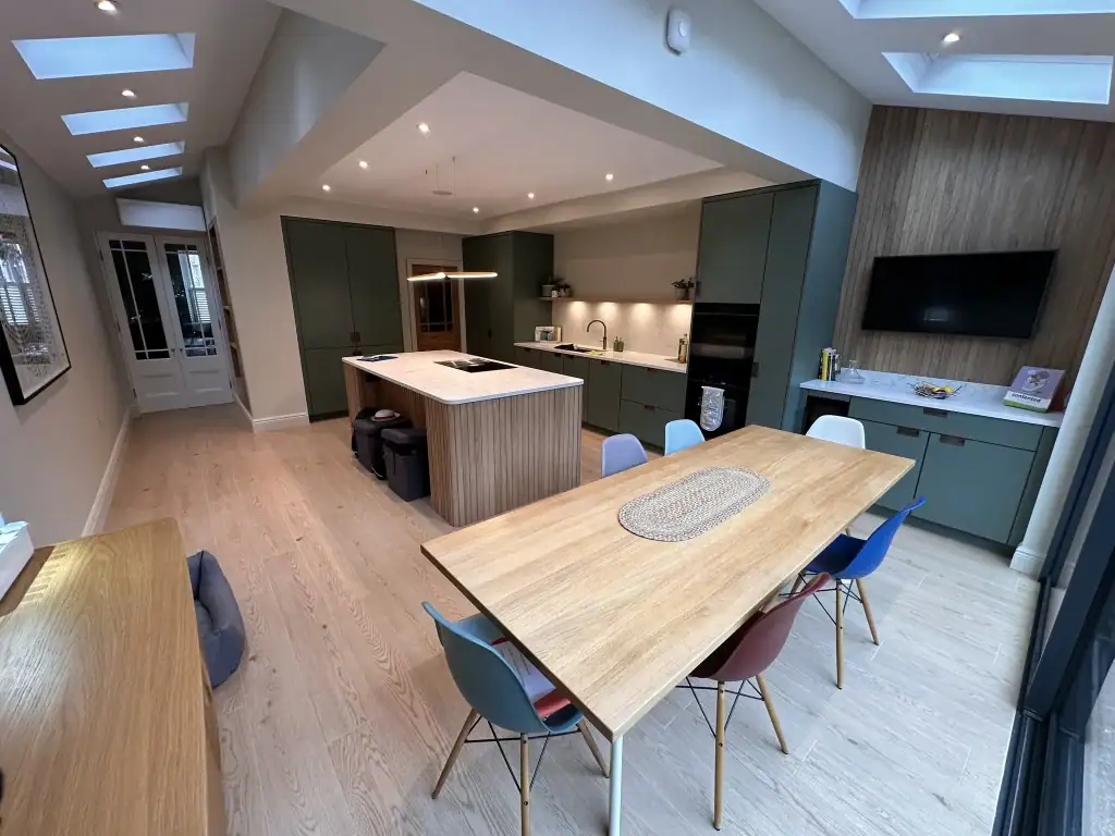 Spacious Clapham kitchen-diner with sage green handleless cabinets, white marble waterfall island, oak dining table with colorful Eames-style chairs, multiple skylights, and light oak flooring in a modern extension