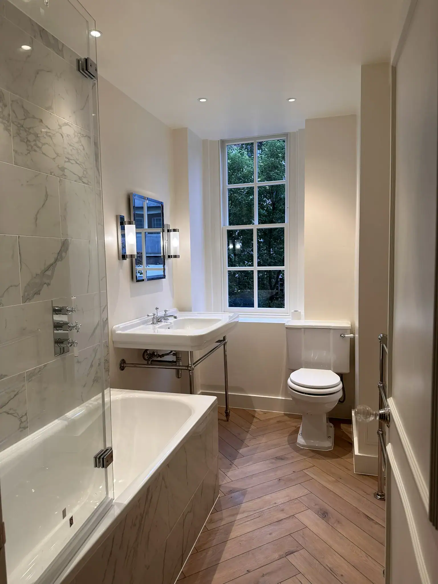 Close-up of ornate white porcelain basin on marble vanity with crosshead taps, adjacent built-in white bathtub, and soft wall sconce lighting in a South Kensington renovation
