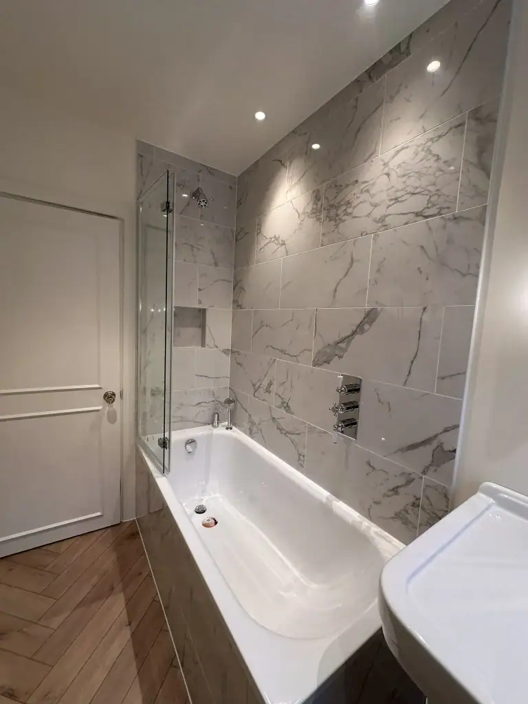 Compact yet luxurious South Kensington bathroom with white marble-tiled shower, built-in bathtub, traditional white vanity basin, herringbone oak flooring, and brass towel radiator