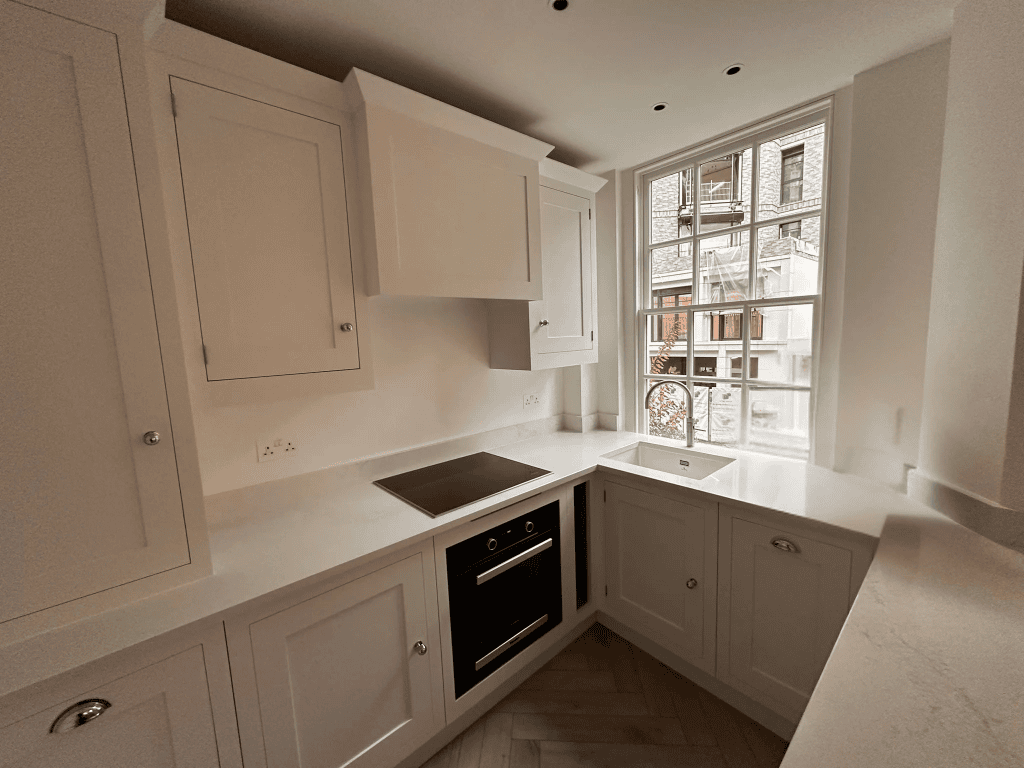 Bright Wimbledon kitchen with white shaker cabinets, matte black integrated oven, white quartz L-shaped worktops, herringbone oak flooring, recessed ceiling lights, and large sash window with urban view